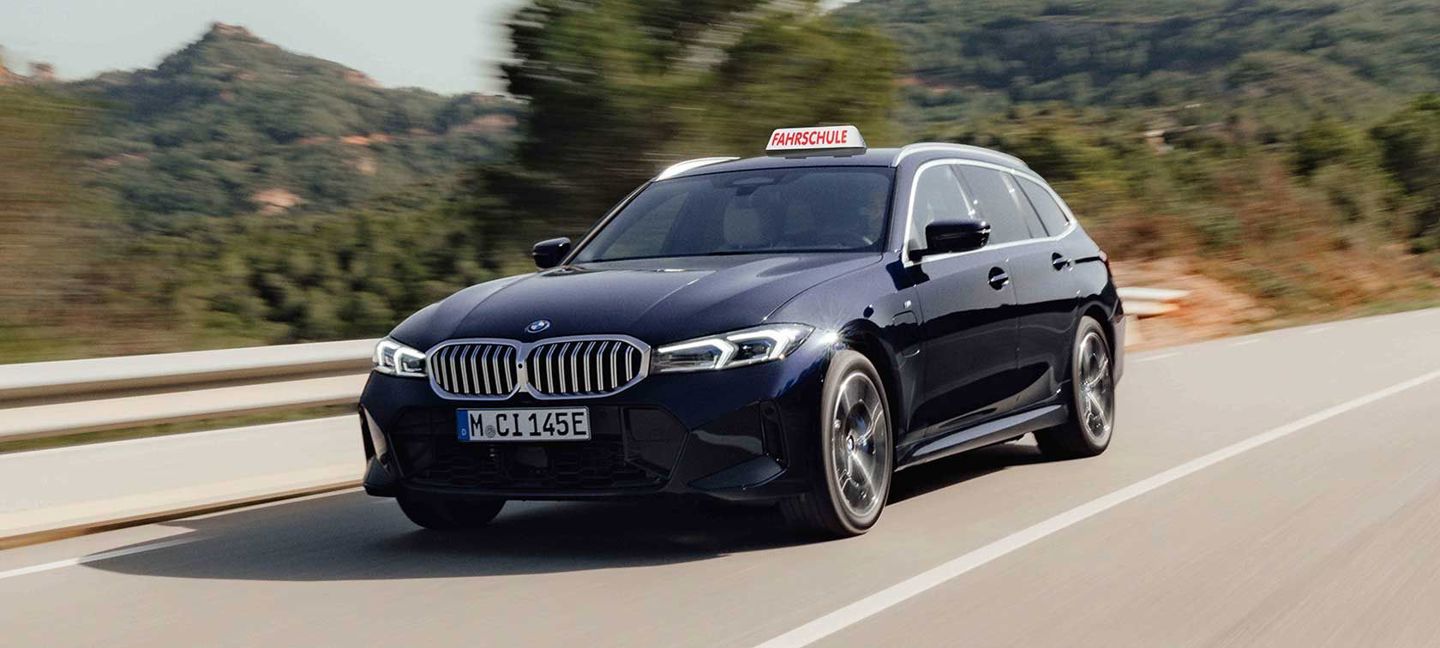 BMW 3er Touring in Dunkelblau mit Fahrschule-Schild fährt auf Landstraße durch grüne Landschaft.“