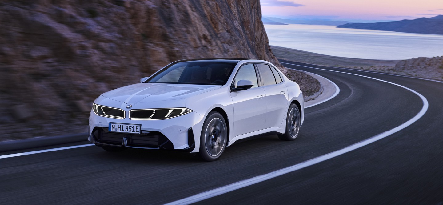 Eine weiße BMW-Limousine fährt auf einer kurvenreichen Küstenstraße entlang felsiger Klippen, im Hintergrund sind das Meer und die Berge zu sehen.