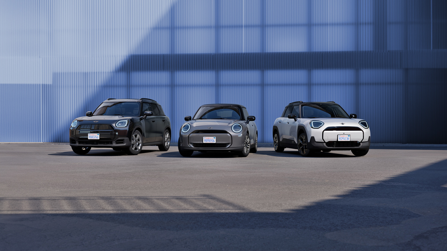 Drei neue MINI Modelle nebeneinander auf einem offenen Platz vor einer blauen Industrie-Fassade: links ein schwarzer MINI Countryman, in der Mitte ein grauer vollelektrischer MINI Cooper und rechts ein weißer MINI Aceman. Die Szene ist modern inszeniert mit klarem Licht-Schatten-Spiel.