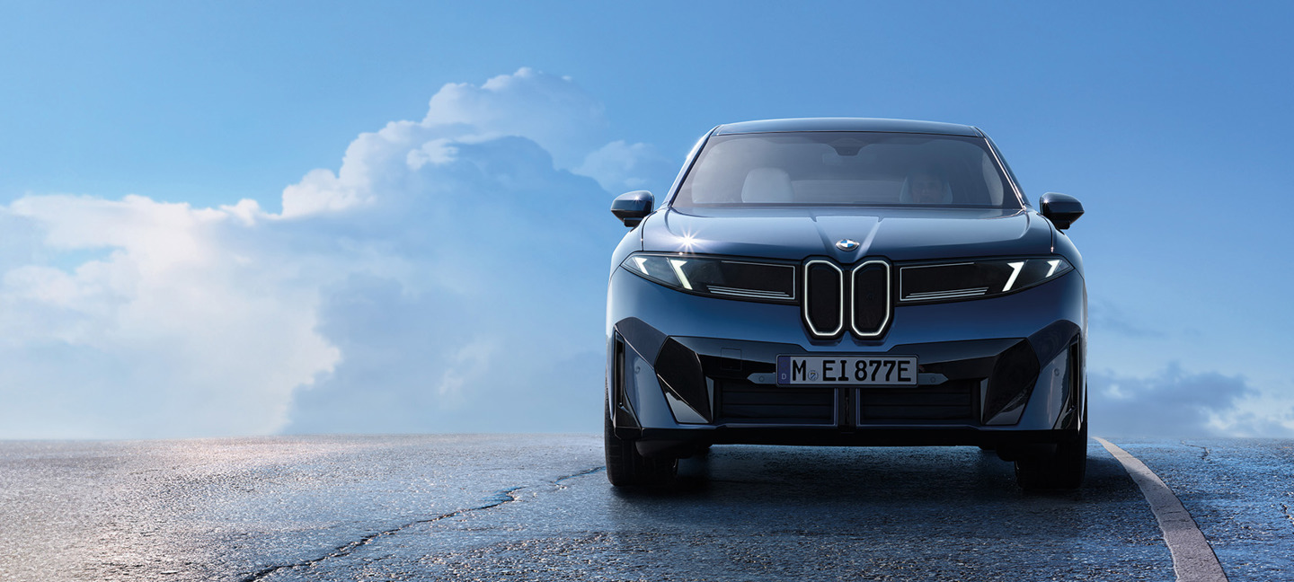 Frontansicht des vollelektrischen BMW iX3 in dunklem Blau, aufgenommen auf freier Straße vor blauem Himmel mit Wolken.