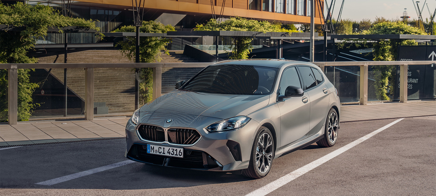 Ein silberner BMW 1er mit Schrägheck ist auf einer Straße vor einem modernen, begrünten Gebäude geparkt.