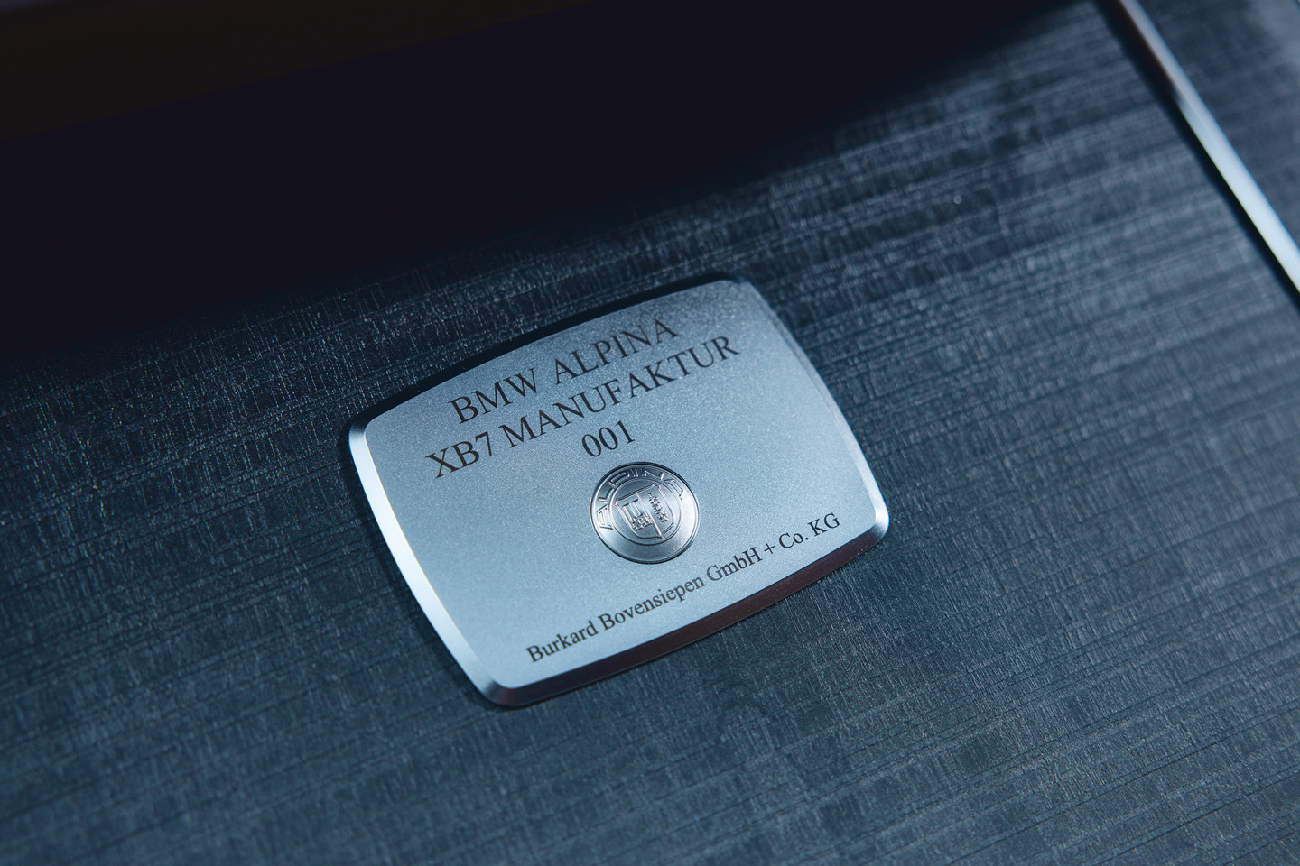 „Detailaufnahme einer silbernen BMW ALPINA Manufakturplakette mit der Gravur 'BMW ALPINA XB7 MANUFAKTUR 001' und dem ALPINA-Logo, montiert auf einer edlen Holz- oder Metalloberfläche im Innenraum.“