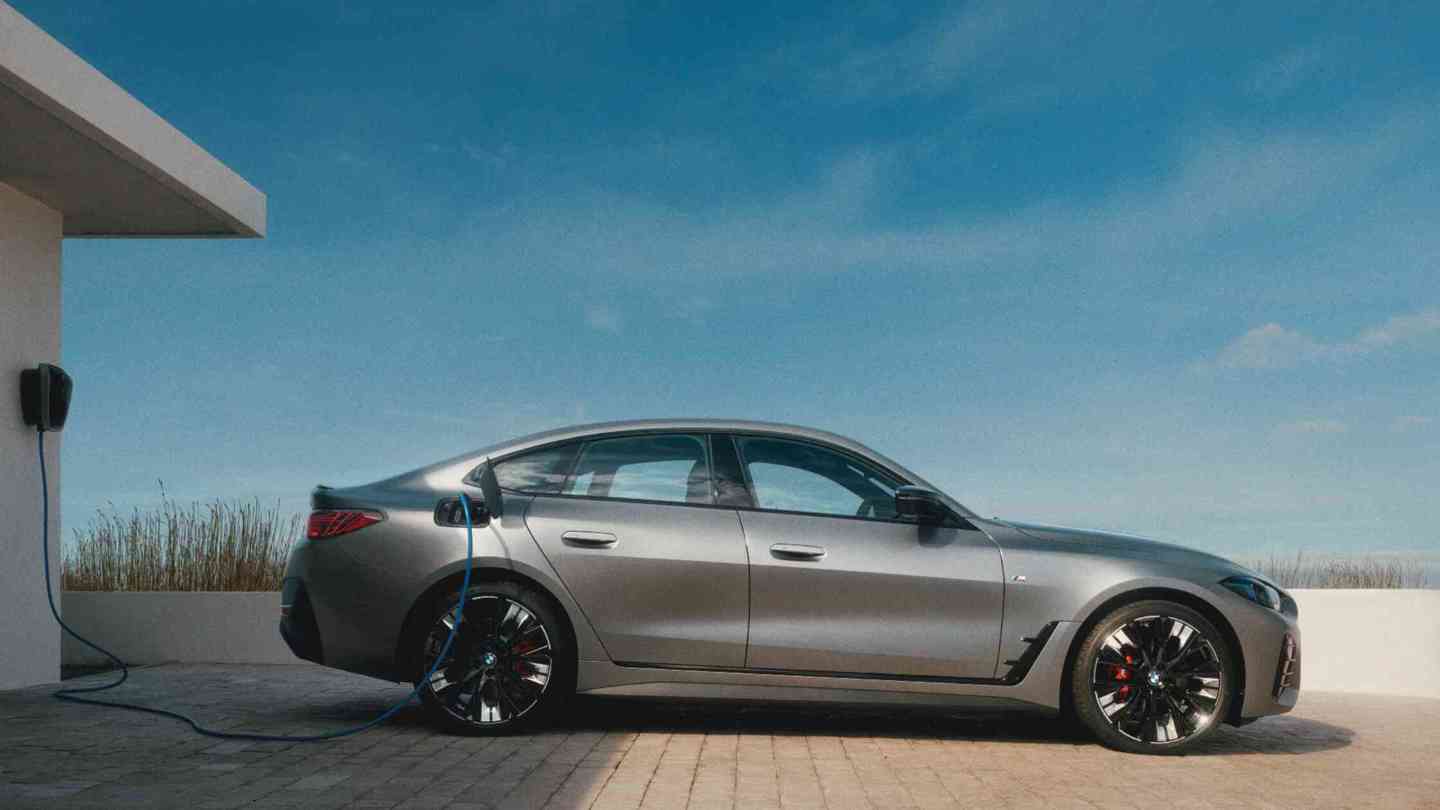 „Ein BMW i4 in mattem Grau steht an einer modernen Hauswand und wird über eine Wallbox geladen. Der Himmel ist klar und blau – die Szene vermittelt nachhaltige Mobilität und stilvolles Design in einem zeitgemäßen Lebensumfeld.“