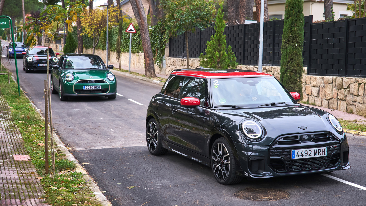 „Mehrere MINI Fahrzeuge fahren hintereinander auf einer Straße. Vorne ein schwarzer MINI mit rotem Dach und roten Spiegelkappen, gefolgt von weiteren MINI Modellen in verschiedenen Farben.“