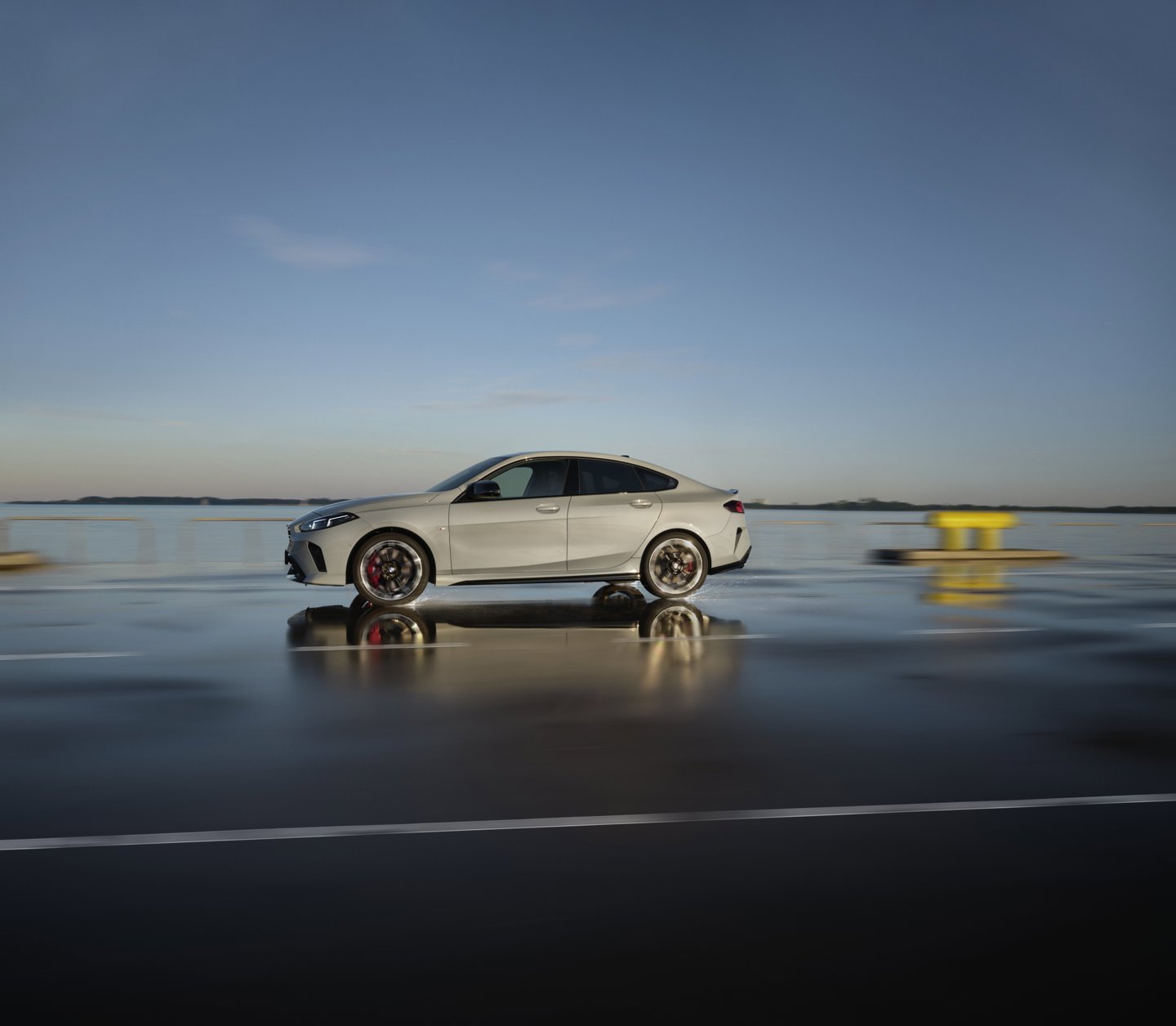 Eine silberne Limousine gleitet bei klarem Himmel auf einer nassen Straße neben einem Gewässer entlang, wobei ihre Bewegung eine auffällige Reflexion auf der glänzenden Oberfläche erzeugt.
