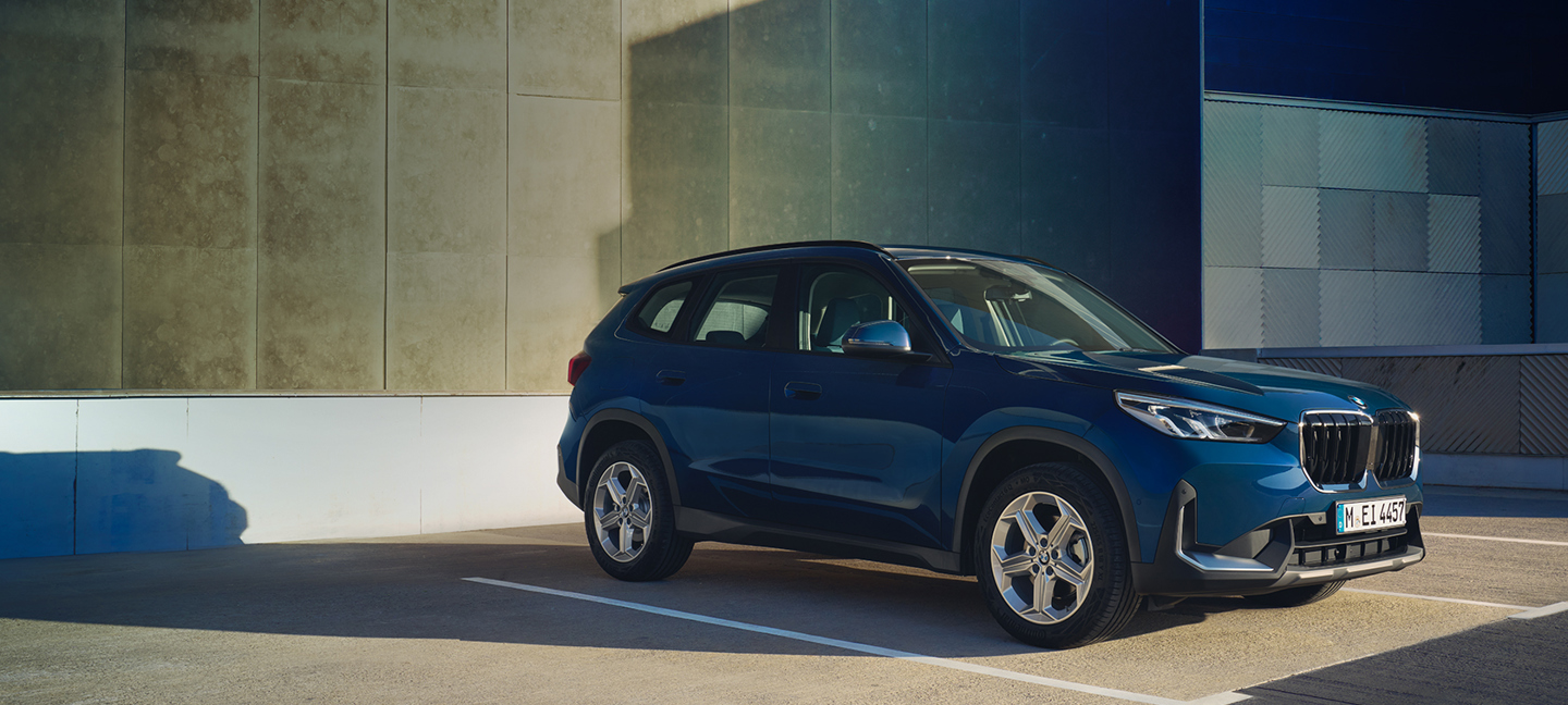 Ein blauer BMW X1 steht auf einem Parkplatz im Freien neben einem Gebäude mit einer modernen, strukturierten Fassade.