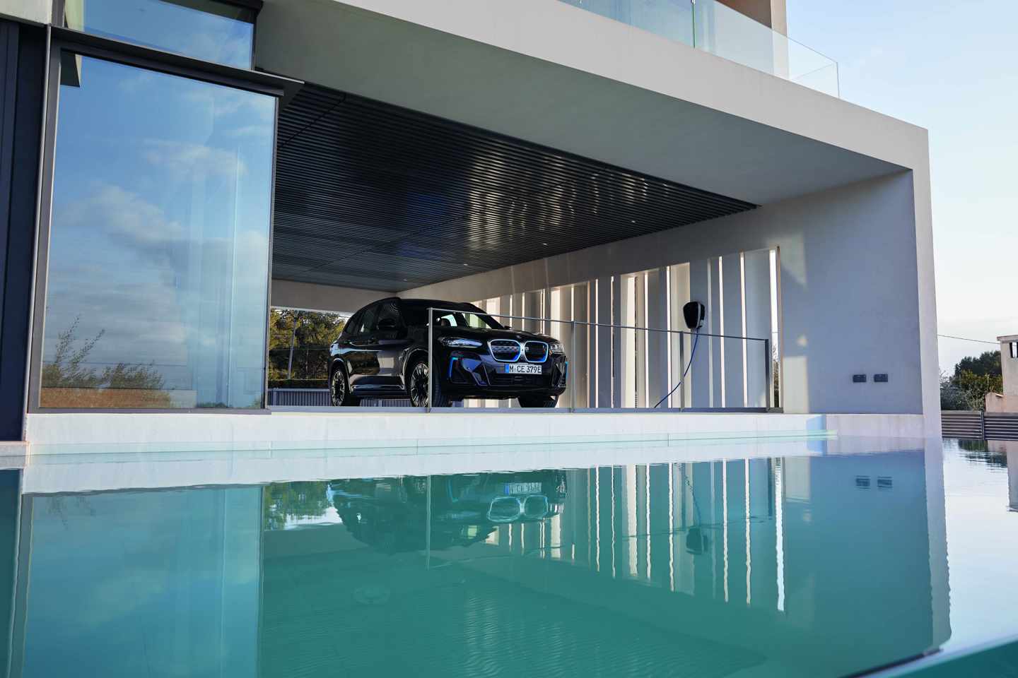 Ein schwarzer Geländewagen parkt in einer modernen, offenen Garage neben einem Swimmingpool mit spiegelnder Wasseroberfläche.