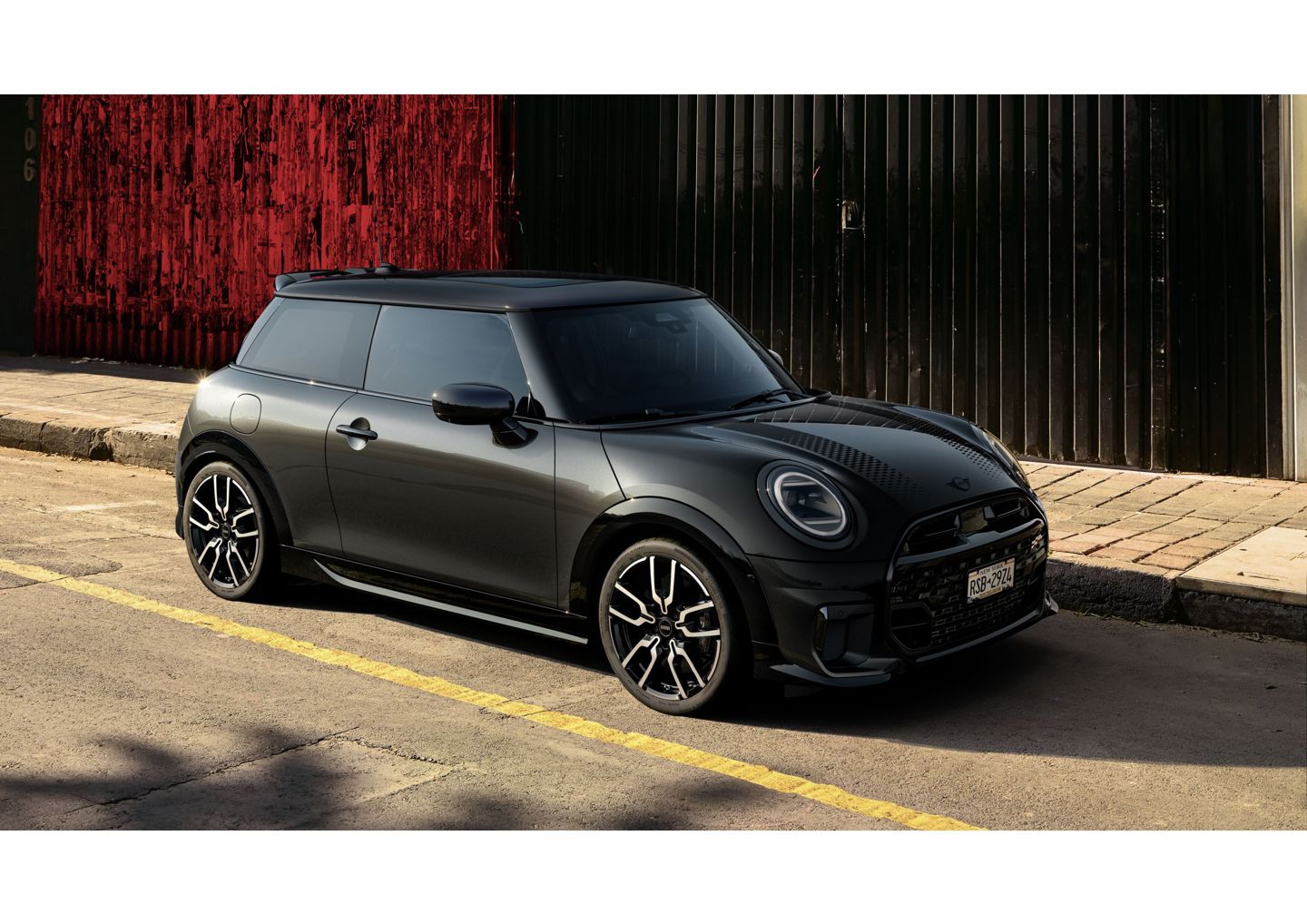Ein schwarzer MINI 3-Türer mit sportlichem Design steht am Straßenrand vor einer schwarz-roten Industriewand in urbaner Umgebung bei Tageslicht.