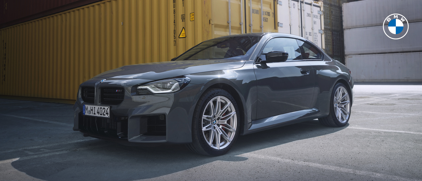 Ein graues BMW-Coupé, das in einem Industriegebiet in der Nähe von gestapelten Frachtcontainern geparkt ist, mit dem BMW-Logo in der rechten oberen Ecke.