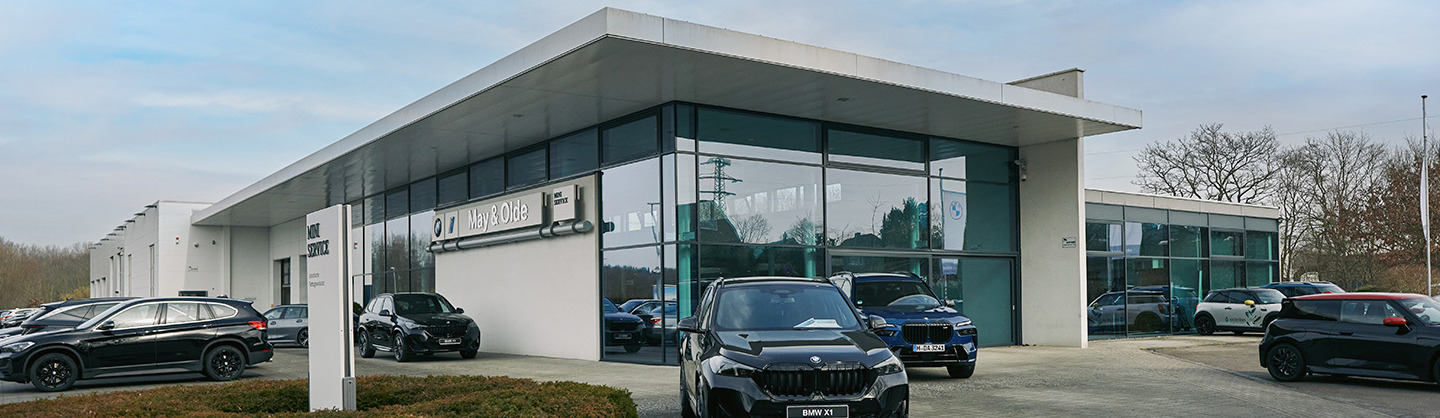 „Außenansicht des Autohauses May & Olde mit moderner Glasfassade, BMW und MINI Logos sowie mehreren schwarzen BMW-Fahrzeugen auf dem Parkplatz bei Tageslicht.“