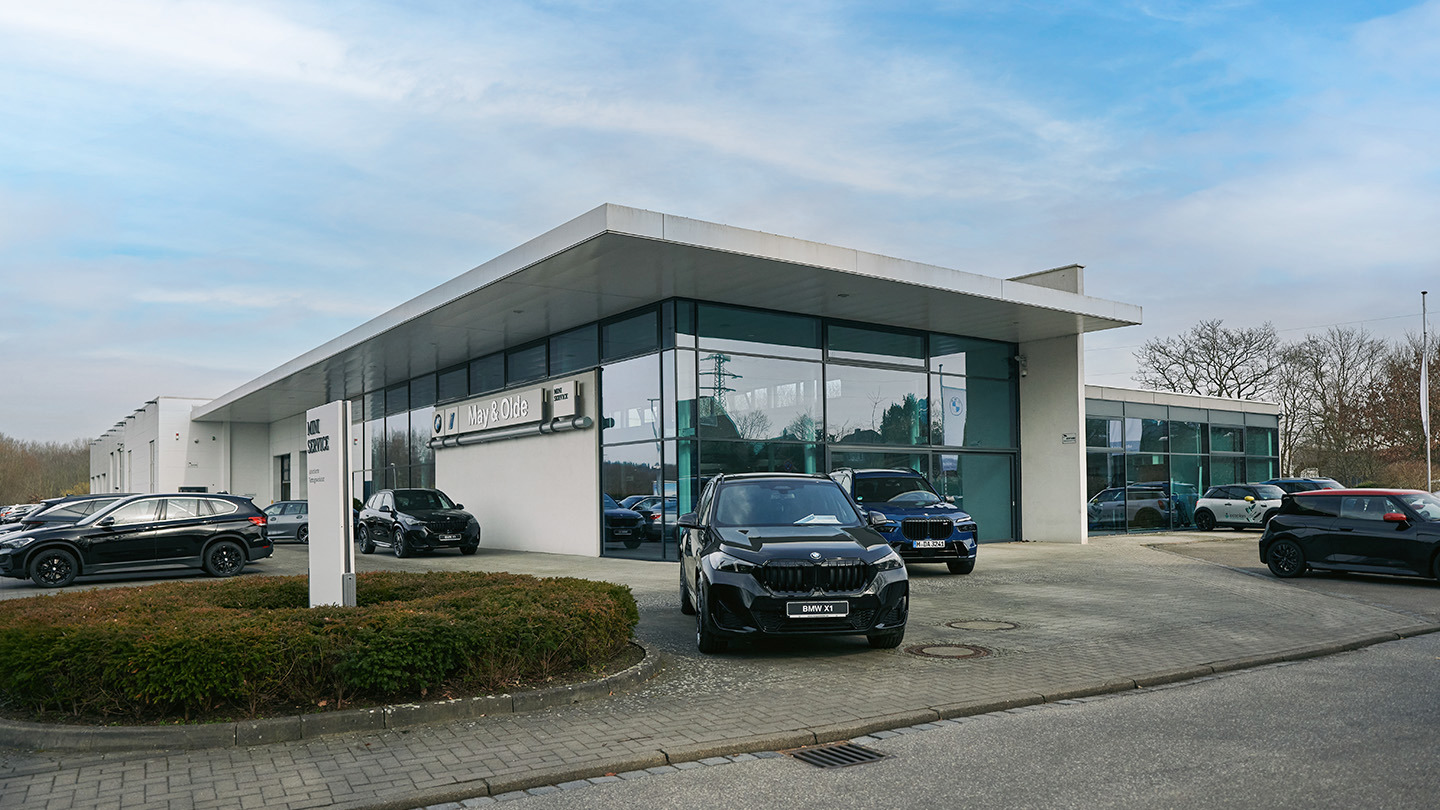 „Außenansicht des Autohauses May & Olde mit moderner Glasfassade, BMW und MINI Logos sowie mehreren schwarzen BMW-Fahrzeugen auf dem Parkplatz bei Tageslicht.“