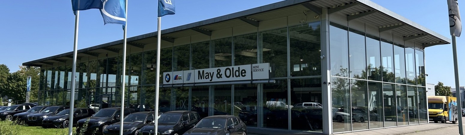 BMW Autohaus May & Olde in Buchholz bei sonnigem Wetter, mit Glasfassade, blauen BMW-Flaggen und mehreren schwarzen BMW-Fahrzeugen vor dem Gebäude aufgereiht .