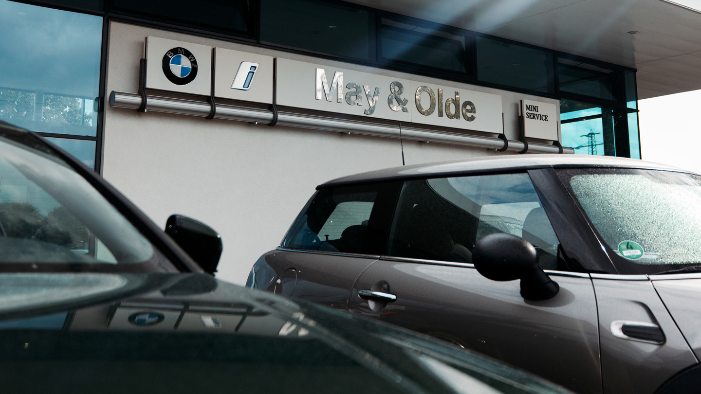 „Außenansicht eines BMW und MINI Autohauses von May & Olde mit sichtbarem Marken-Schriftzug über dem Eingang. Im Vordergrund stehen ein MINI und ein BMW-Fahrzeug, die leicht von Regentropfen benetzt sind. Die Fassade ist modern mit Glasfront und reflektiert das Tageslicht.“