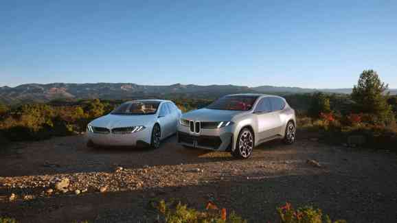 „Zwei futuristische BMW Fahrzeuge stehen nebeneinander auf einem Schotterweg inmitten einer malerischen Landschaft. Links ein weißes, flaches Konzeptfahrzeug in Limousinenform, rechts ein silberner SUV mit markanter Front. Im Hintergrund sind grüne Hügel, Wälder und eine Gebirgskette unter blauem Himmel zu sehen.“