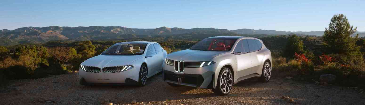 „Zwei futuristische BMW Fahrzeuge stehen nebeneinander auf einem Schotterweg inmitten einer malerischen Landschaft. Links ein weißes, flaches Konzeptfahrzeug in Limousinenform, rechts ein silberner SUV mit markanter Front. Im Hintergrund sind grüne Hügel, Wälder und eine Gebirgskette unter blauem Himmel zu sehen.“
