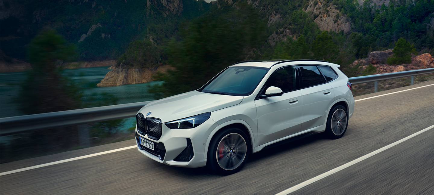 BMW iX1 in Weiß fährt dynamisch auf einer kurvigen Landstraße entlang eines Sees – elektrisches SUV mit sportlich-elegantem Design in Bewegung.