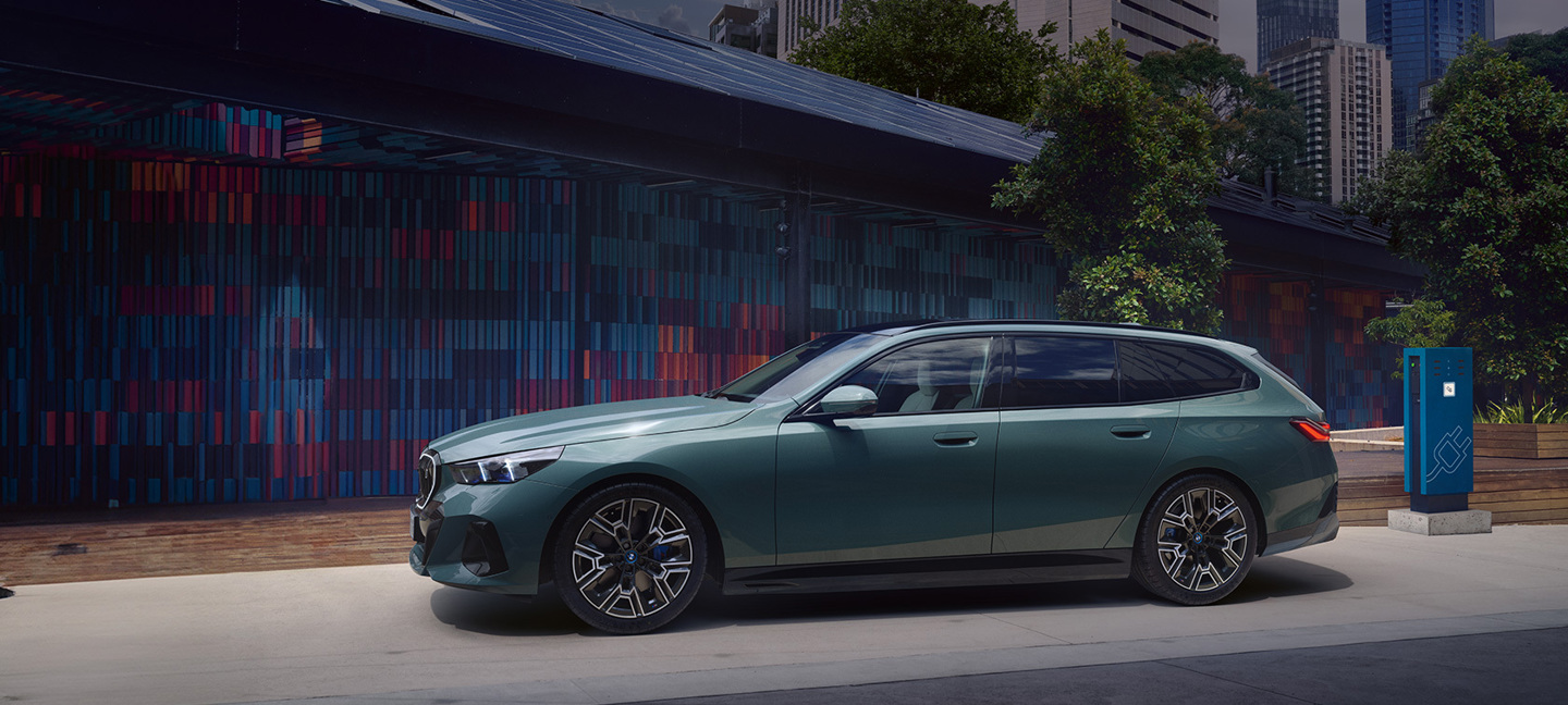 BMW i5 Touring in Grün seitlich an einer E-Ladesäule in urbaner Umgebung geparkt. Das elegante Elektrofahrzeug steht vor einer modernen Wand mit farbenfrohen Paneelen, umgeben von Bäumen und Hochhäusern im Hintergrund.