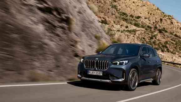 „Ein dunkler BMW X1 fährt dynamisch durch eine kurvige Gebirgsstraße. Im Hintergrund sind felsige Hänge mit spärlicher Vegetation und ein klarer blauer Himmel zu sehen.“