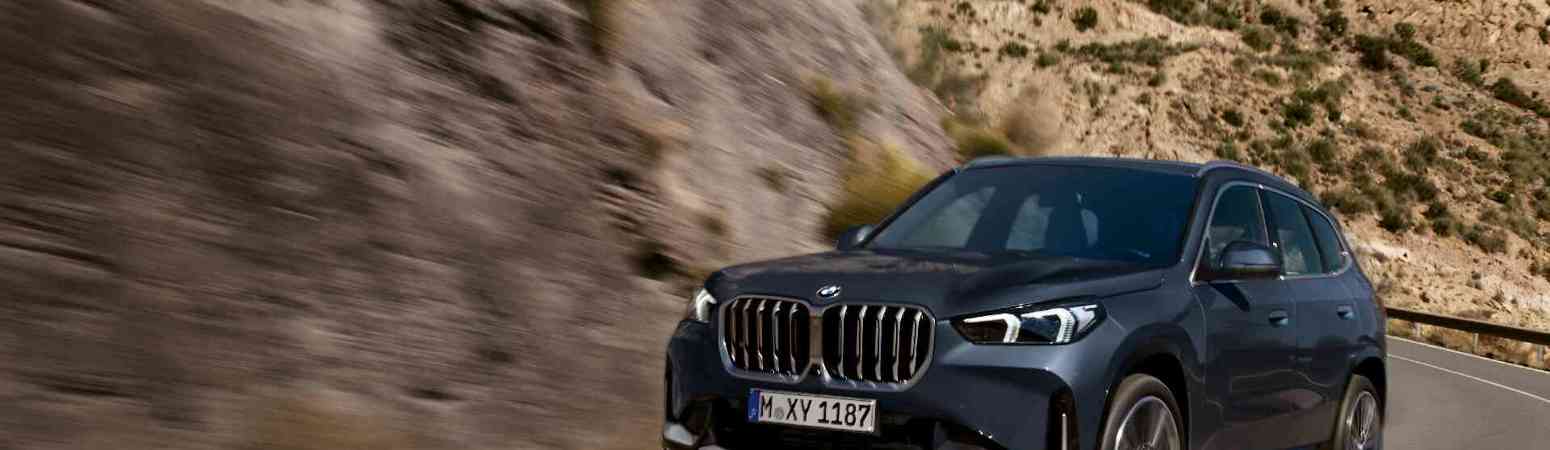„Ein dunkler BMW X1 fährt dynamisch durch eine kurvige Gebirgsstraße. Im Hintergrund sind felsige Hänge mit spärlicher Vegetation und ein klarer blauer Himmel zu sehen.“