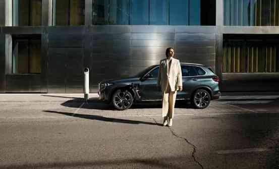 „Eine Person im hellen Hosenanzug steht vor einem dunkelgrauen BMW SUV, der an einer Ladesäule angeschlossen ist. Im Hintergrund ist eine moderne Glas- und Metallfassade zu sehen.“