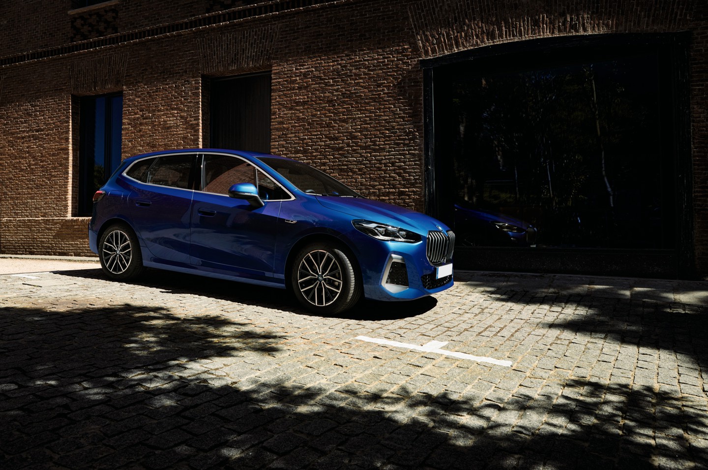 Ein blauer BMW 2er Active Tourer steht auf einer gepflasterten Straße vor einer modernen Backsteinfassade. Das Fahrzeug ist seitlich zu sehen, mit markanter Front und großen Felgen. Im Hintergrund befinden sich dunkle Fensterflächen, die das Auto spiegeln. Sonnenlicht wirft Schatten von Bäumen auf die Straße und das Fahrzeug.