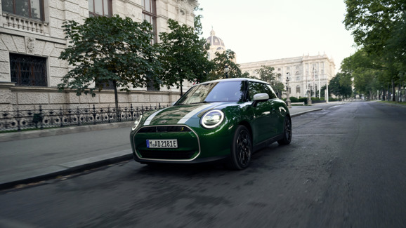 Grüner MINI Cooper SE in Fahrt durch eine historische Innenstadtallee, mit markanten weißen Rennstreifen und modernem LED-Tagfahrlicht