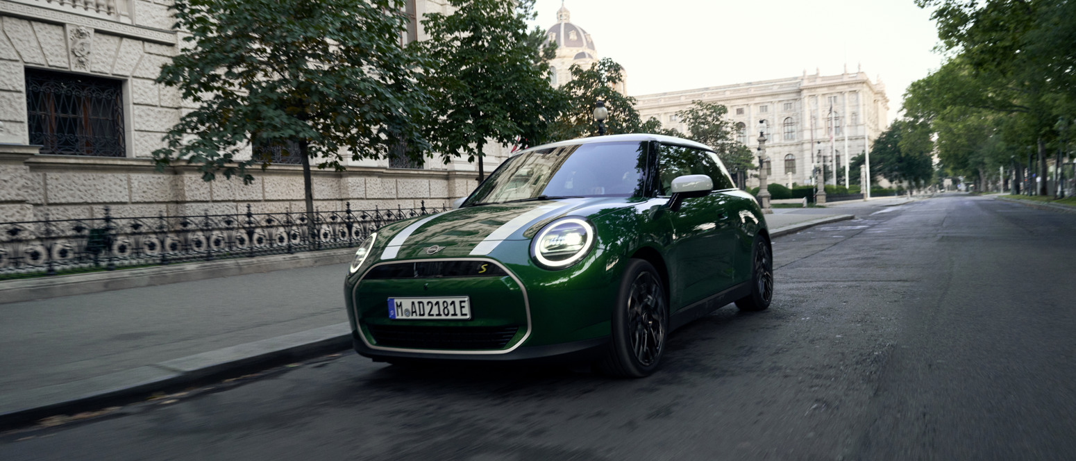 Grüner MINI Cooper SE in Fahrt durch eine historische Innenstadtallee, mit markanten weißen Rennstreifen und modernem LED-Tagfahrlicht