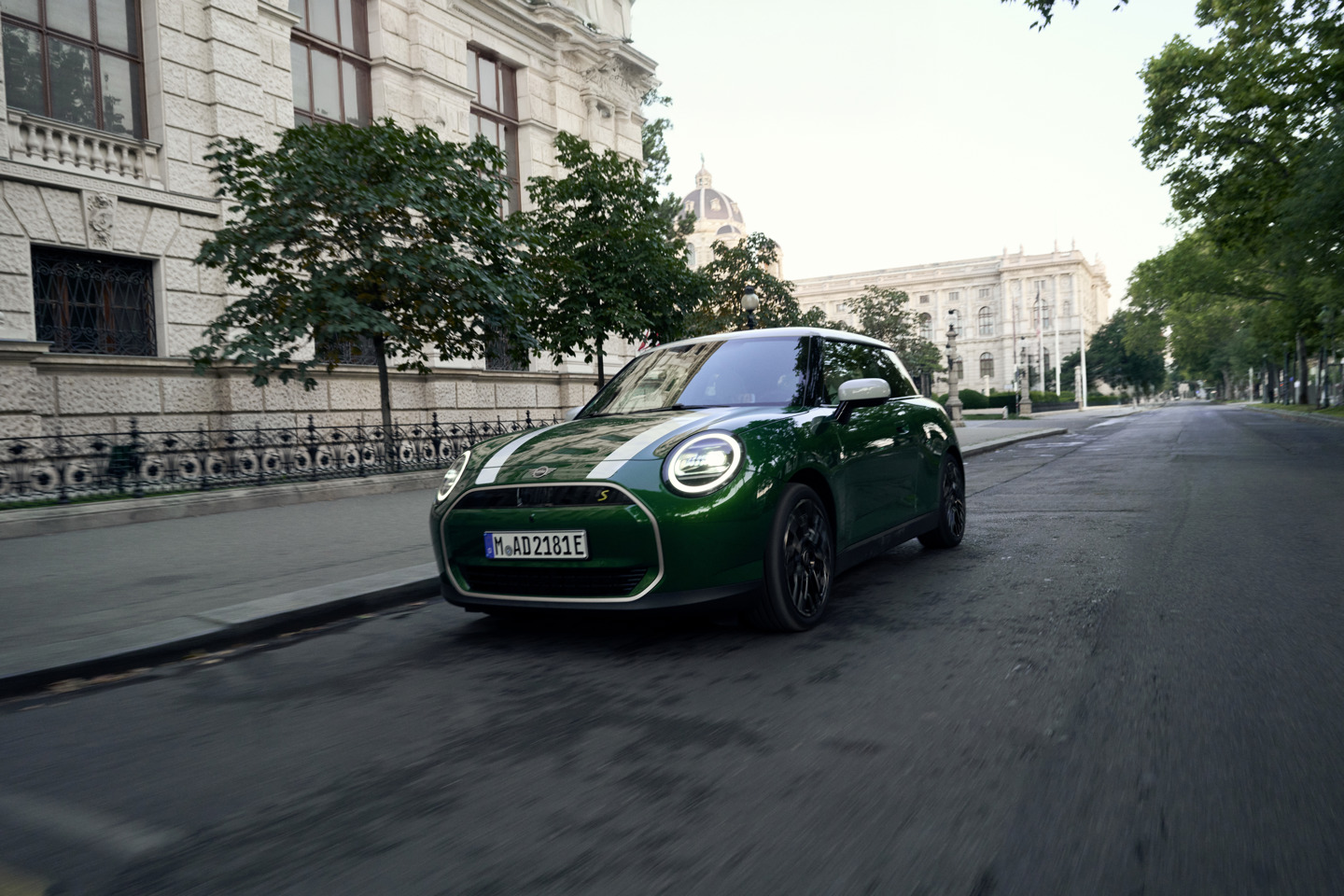 Grüner MINI Cooper SE in Fahrt durch eine historische Innenstadtallee, mit markanten weißen Rennstreifen und modernem LED-Tagfahrlicht