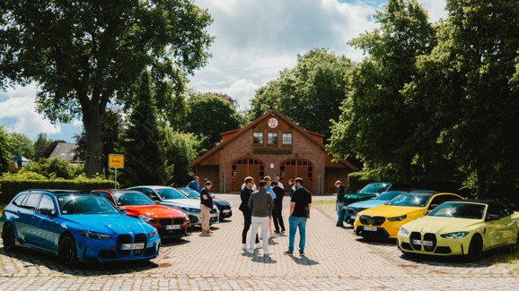 An einem sonnigen Tag steht eine Gruppe von Menschen in einem Innenhof vor einem Backsteingebäude, in dem mehrere BMW M-Fahrzeuge geparkt sind.