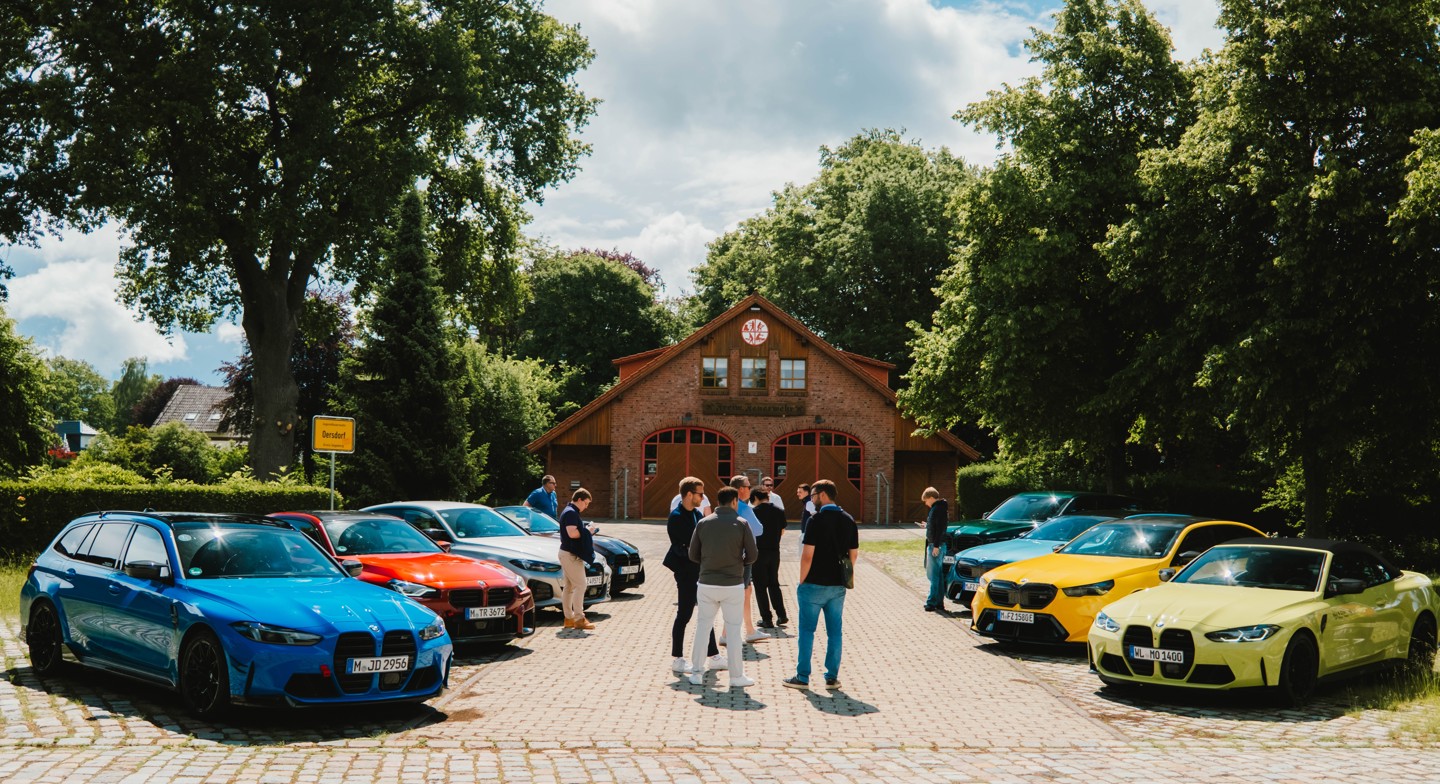An einem sonnigen Tag steht eine Gruppe von Menschen in einem Innenhof vor einem Backsteingebäude, in dem mehrere BMW M-Fahrzeuge geparkt sind.