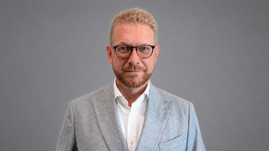 Ein Mann mit kurzen blonden Haaren, Bart und Brille, der einen hellgrauen Anzug und ein weißes Hemd trägt und vor einem schlichten grauen Hintergrund steht.