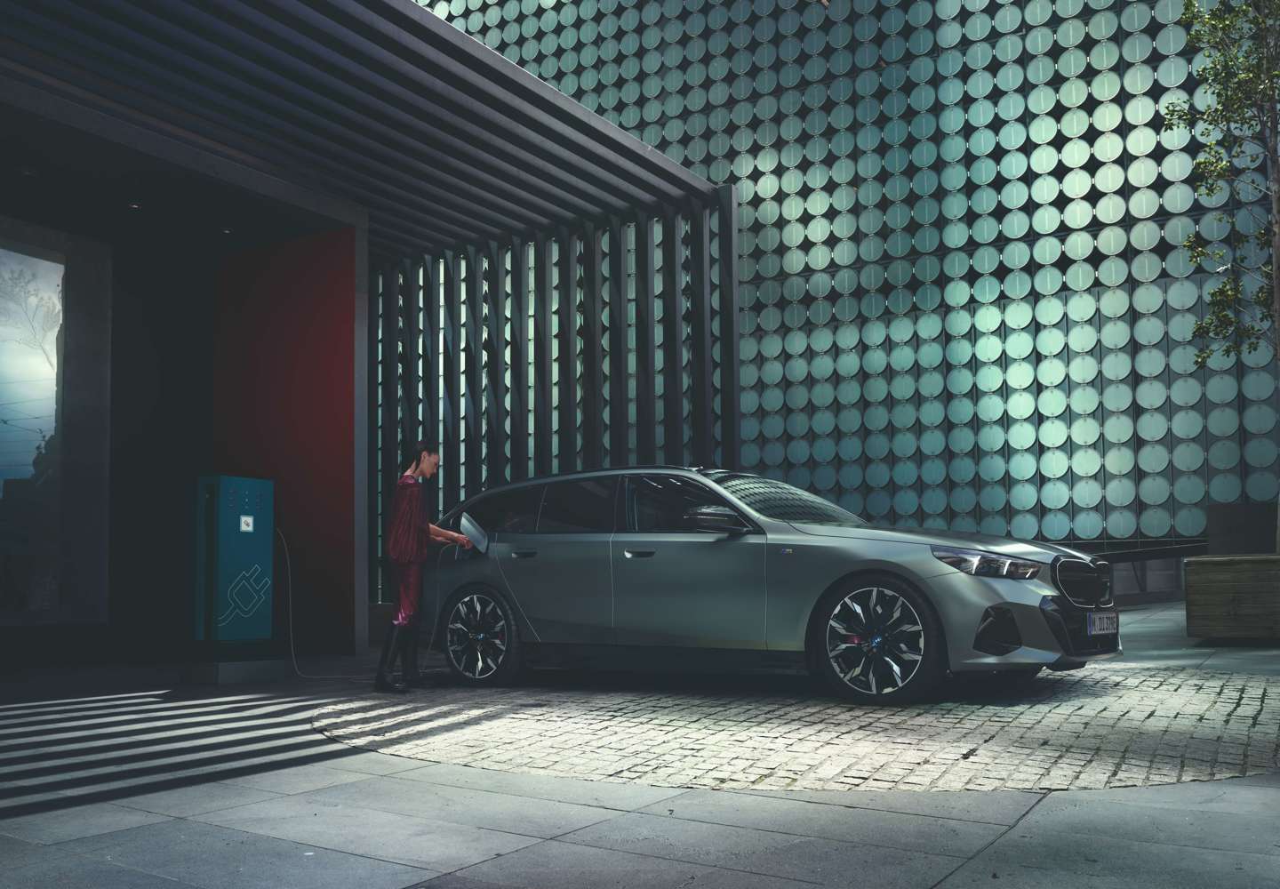 „Eine Frau lädt ein modernes, grau lackiertes BMW Elektrofahrzeug an einer öffentlichen Ladestation auf. Die Szene spielt sich in einem urbanen Umfeld mit moderner Architektur und reflektierender Fassadenstruktur ab. Das Licht erzeugt ein stimmungsvolles Spiel aus Schatten und Glanz.“