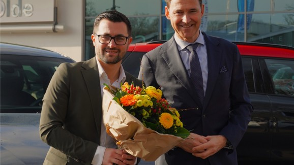 „Zwei Männer in Anzügen stehen vor einem Autohaus. Der Mann links trägt eine Brille und hält einen bunten Blumenstrauß, der Mann rechts legt ihm die Hand auf den Arm. Beide lächeln in die Kamera. Im Hintergrund sind BMW- und MINI-Fahrzeuge zu sehen.“