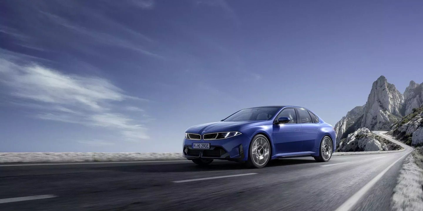 Ein blauer BMW fährt unter klarem Himmel eine menschenleere Bergstraße entlang, im Hintergrund ragen schroffe Felsgipfel empor.