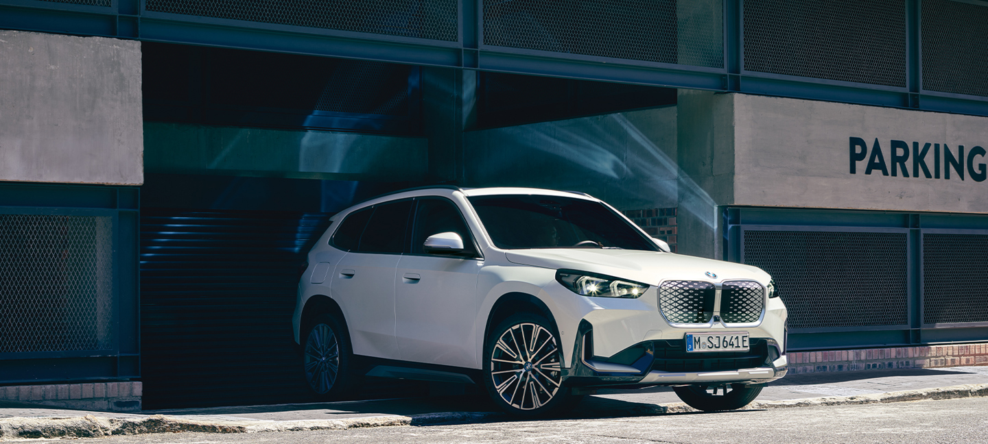 Ein weißer BMW iX1 steht vor der Ausfahrt eines Parkhauses im Sonnenlicht. Das moderne Elektro-SUV präsentiert sich mit markanter Front und beleuchteten Nieren.
