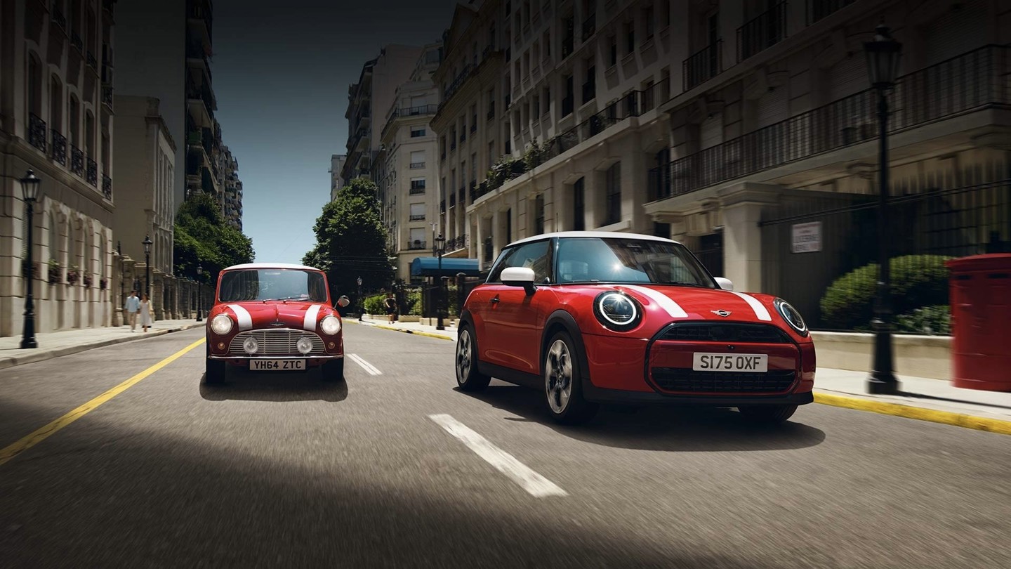 Zwei rote MINI Fahrzeuge, ein klassischer MINI Oldtimer und ein moderner MINI Cooper, fahren nebeneinander auf einer städtischen Straße mit historischer Architektur im Hintergrund, dynamische Fahrszene mit Fokus auf Designentwicklung, Tradition und modernes Fahrgefühl ohne eingeblendeten Werbetext
