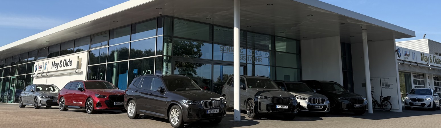 BMW Autohaus May & Olde in Halstenbek mit moderner Glasfassade und mehreren ausgestellten BMW Fahrzeugen vor dem Eingang bei sonnigem Wetter.