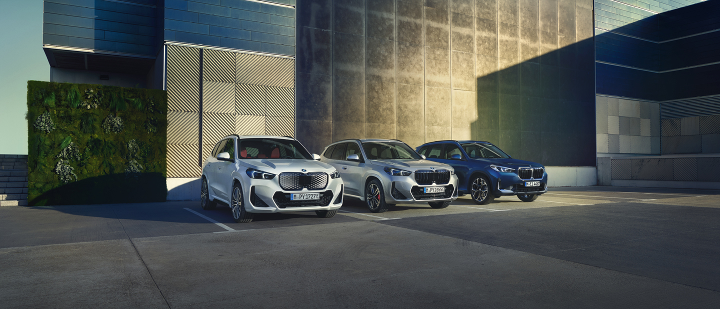 Drei BMW X1 Modelle nebeneinander vor einer modernen Gebäudefassade: links ein weißer BMW X1 xDrive25e Plug-in-Hybrid, in der Mitte ein silberner BMW X1 mit sportlicher M-Ausstattung und rechts ein blauer vollelektrischer BMW iX1. Im Hintergrund ein blauer Kreis mit dem Schriftzug „Alle Angebote inkl. Wartung und Reparatur.“