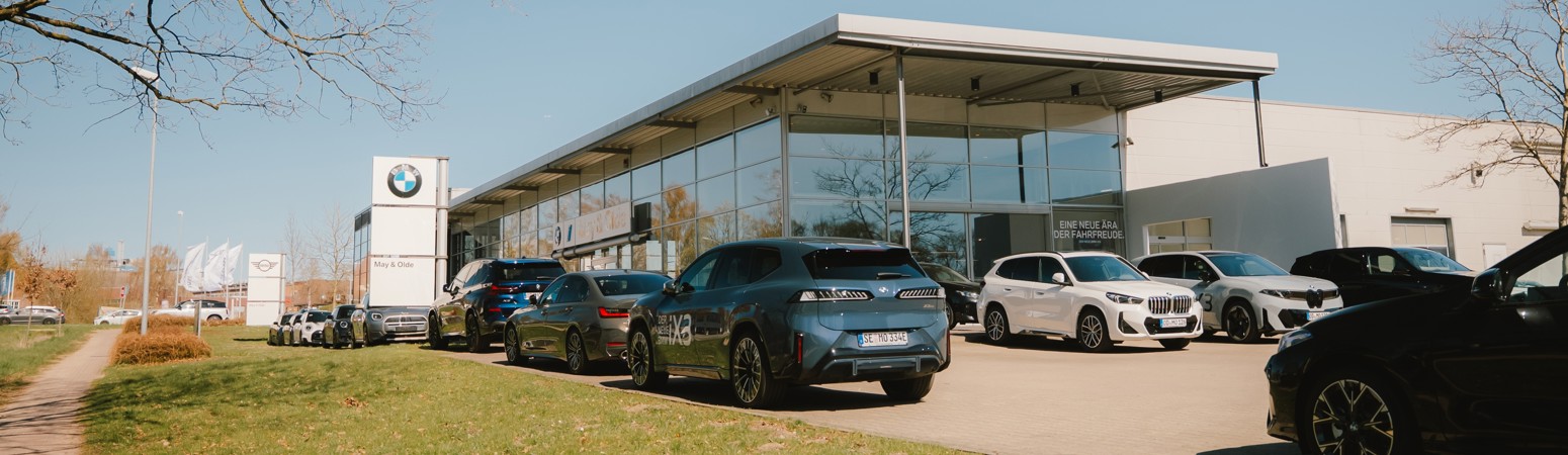 Außenansicht des Autohauses May & Olde am Standort Ahrensburg mit moderner Glasfassade und BMW Branding, mehrere BMW Fahrzeuge auf dem Parkplatz vor dem Gebäude, Grünfläche und Gehweg im Vordergrund sowie klare Himmelsszene, Standortaufnahme mit Fokus auf Ausstellung und Erreichbarkeit
