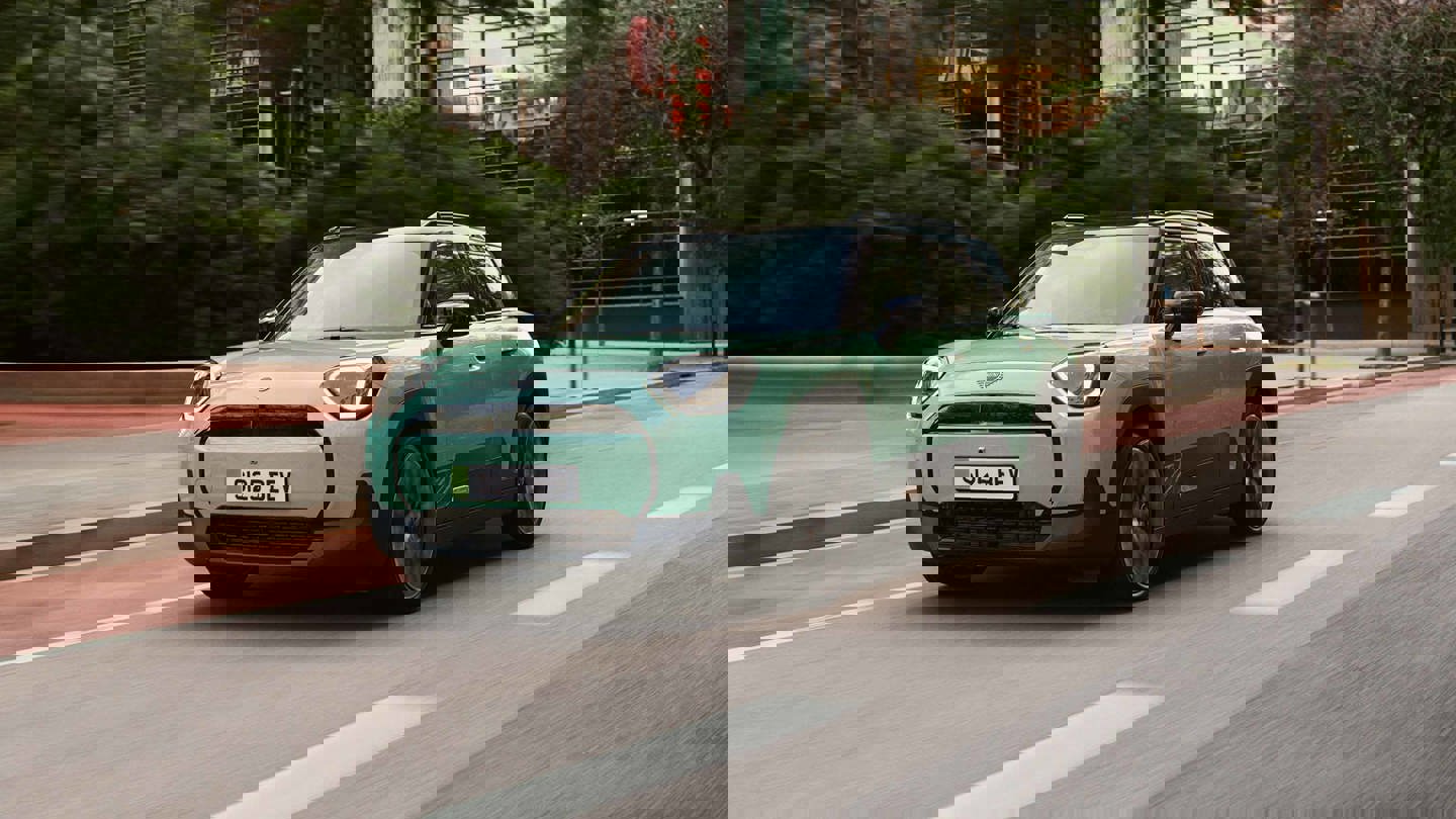 MINI Aceman in grün als kompakter SUV in dynamischer Fahrszene auf einer urbanen Straße, unterwegs durch eine moderne Stadt mit Bäumen und Gebäuden im Hintergrund, Fokus auf Elektromobilität, Fahrdynamik und urbanen Lifestyle ohne eingeblendeten Werbetext