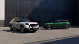 Zwei vollelektrische MINI Modelle vor einer dunklen Gebäudefassade: links ein weißer MINI Countryman E mit schwarzem Dach und Münchner Kennzeichen, rechts ein grüner MINI Aceman E in Seitenansicht mit markantem SUV-Design.
