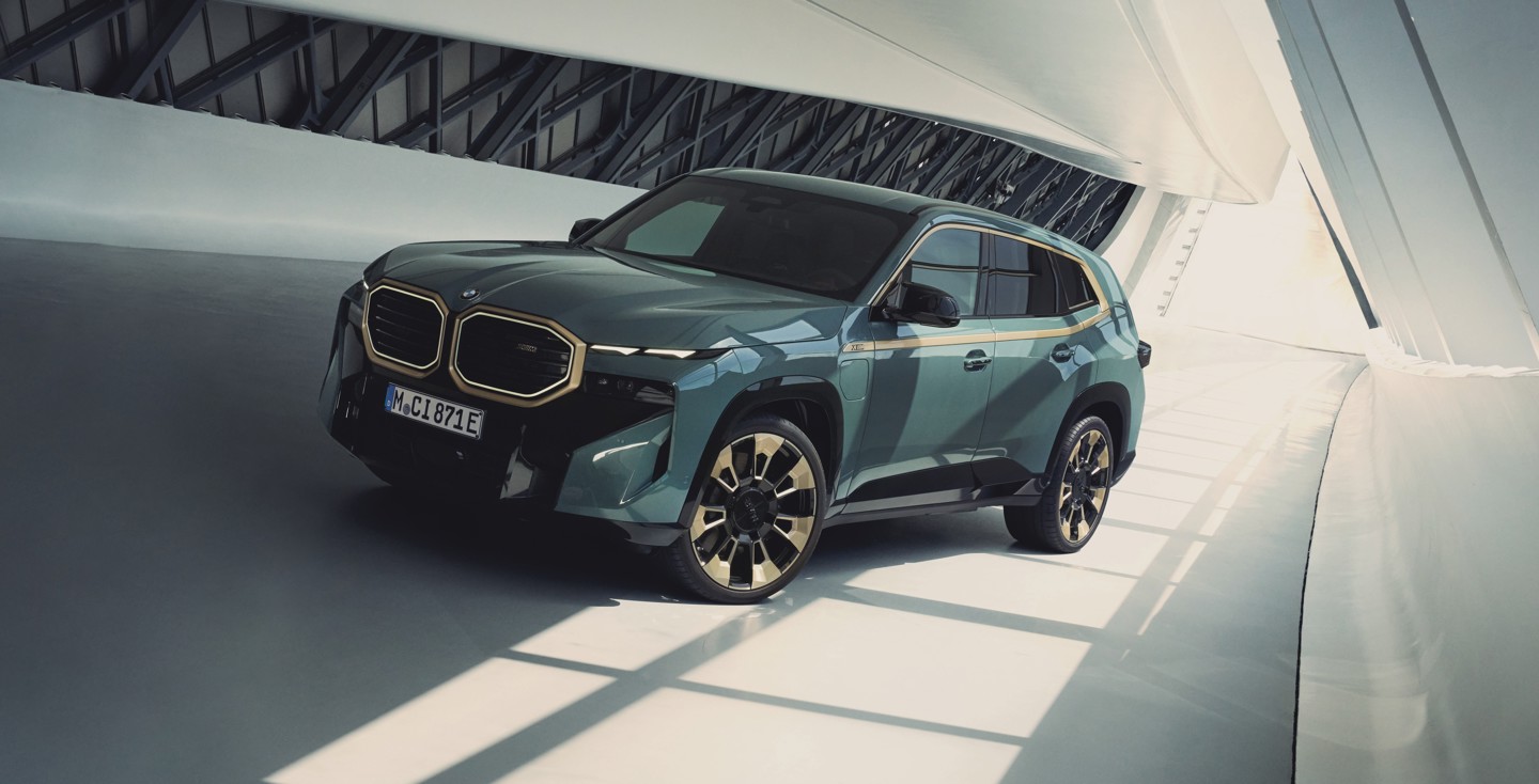 Ein dunkelgrüner BMW XM mit goldenen Verzierungen ist in einem modernen, weißen, lichtdurchfluteten Raum geparkt.