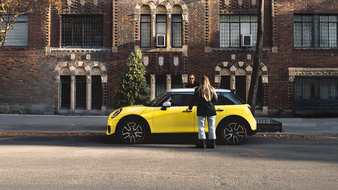 Zwei Personen stehen neben einem gelben Auto MINI Cooper, der auf einer Straße in der Stadt geparkt ist, mit einem Backsteingebäude mit gewölbten Fenstern im Hintergrund.