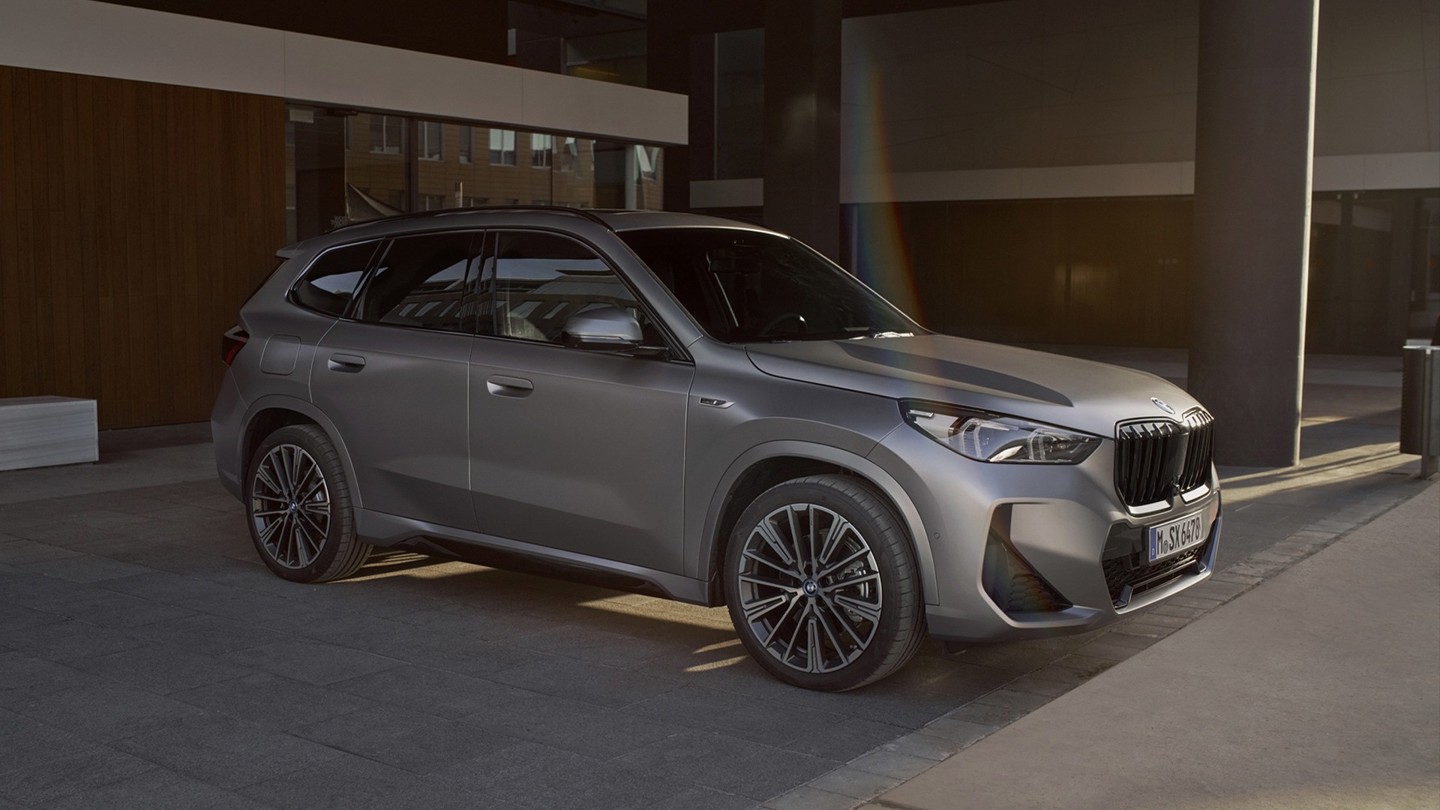 Ein mattgrauer BMW-SUV steht auf einer gepflasterten Fläche in der Nähe eines modernen Gebäudes mit dunklen Säulen und Glasfenstern.