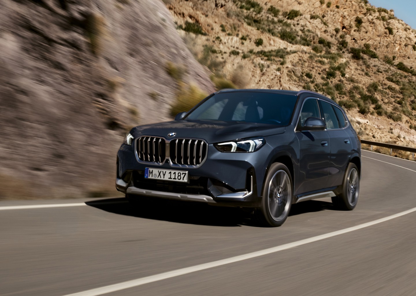Ein dunkelgrauer BMW-SUV fährt auf einer kurvenreichen Bergstraße, im Hintergrund ist felsiges Gelände zu sehen.