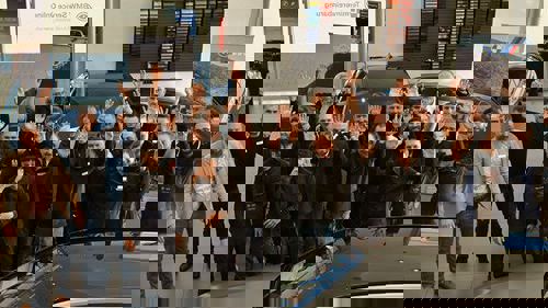 „Ein Team von rund 20 Mitarbeitenden eines Autohauses steht in einer Ausstellungshalle zwischen Fahrzeugen. Alle lächeln und winken in die Kamera. Im Hintergrund sind Farbtafeln, Werbetafeln und große Fenster mit Tageslicht zu sehen.“