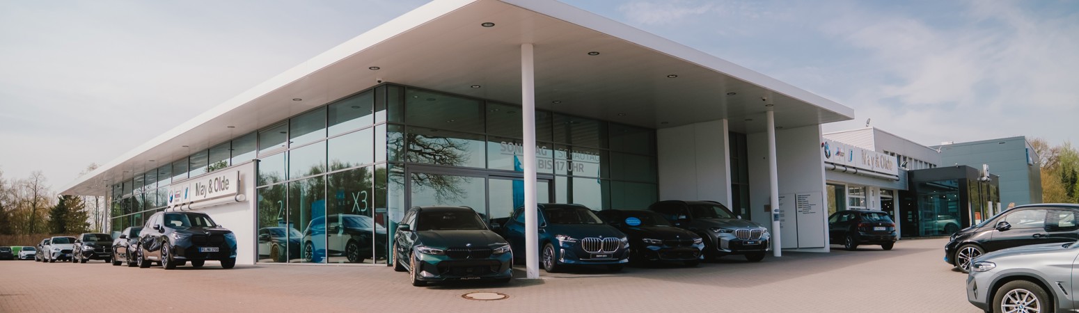 Außenansicht des modernen Autohauses May & Olde am Standort Halstenbek mit großzügiger Glasfassade, ausgestellten BMW Fahrzeugen vor dem Gebäude und weiterem Showroom-Bereich im Hintergrund, weitläufiger Vorplatz mit geparkten Fahrzeugen unter klarem Himmel