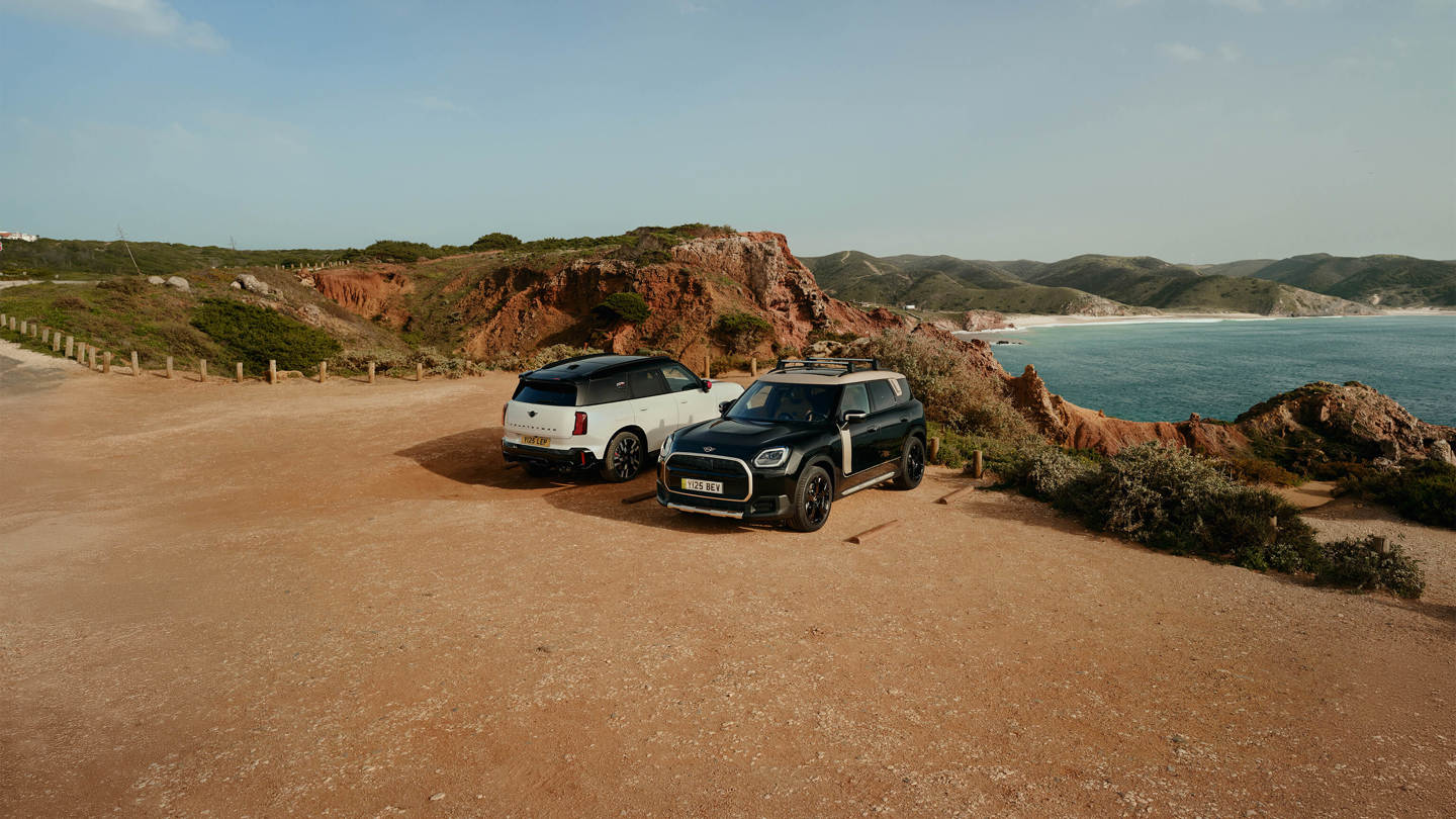 Zwei MINI Fahrzeuge, ein weißer und ein schwarzer vollelektrischer MINI Countryman, geparkt auf einem Aussichtspunkt an einer Küstenlandschaft mit rotem Felsgestein, Meerblick und hügeliger Umgebung im Hintergrund, Offroad-inspirierte Szene mit Fokus auf Abenteuer, Freiheit und modernes Fahrzeugdesign ohne eingeblendeten Werbetext