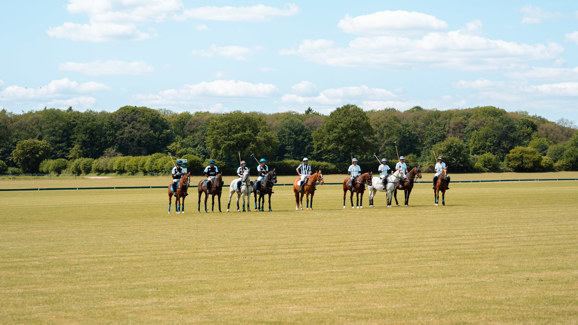 „Zwei Polo-Teams mit jeweils vier Reitern stehen mit ihren Pferden nebeneinander auf einem grünen Spielfeld, im Hintergrund eine Baumreihe unter blauem Himmel mit vereinzelten Wolken.“