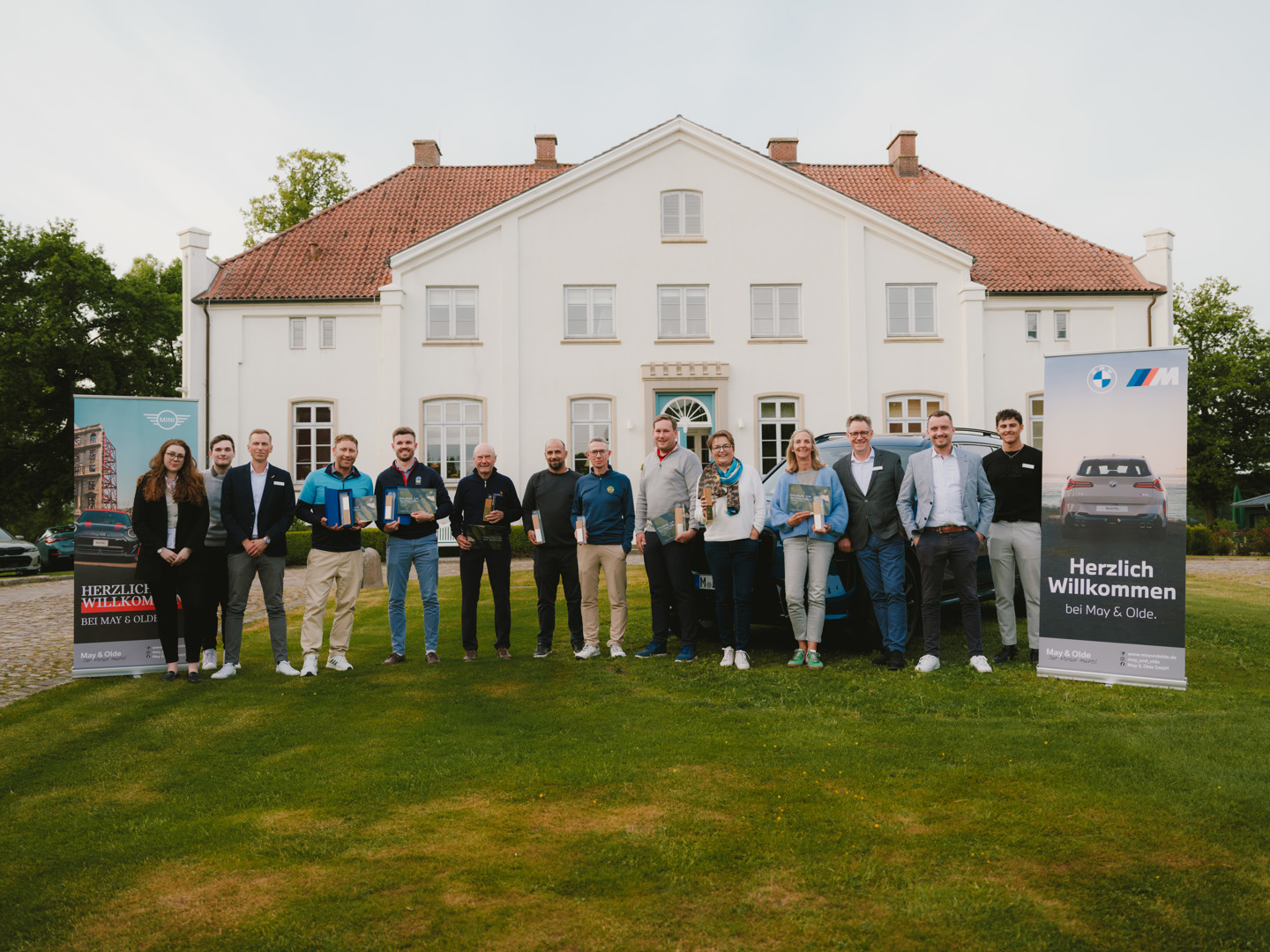 „Gruppenfoto vor einem historischen weißen Herrenhaus mit Gewinnern und Organisatoren des BMW Golf Cup 2024 von May & Olde. Die Personen halten Preise in den Händen, flankiert von zwei Roll-ups mit den Aufschriften ‚Herzlich Willkommen bei May & Olde‘ (BMW & MINI).“